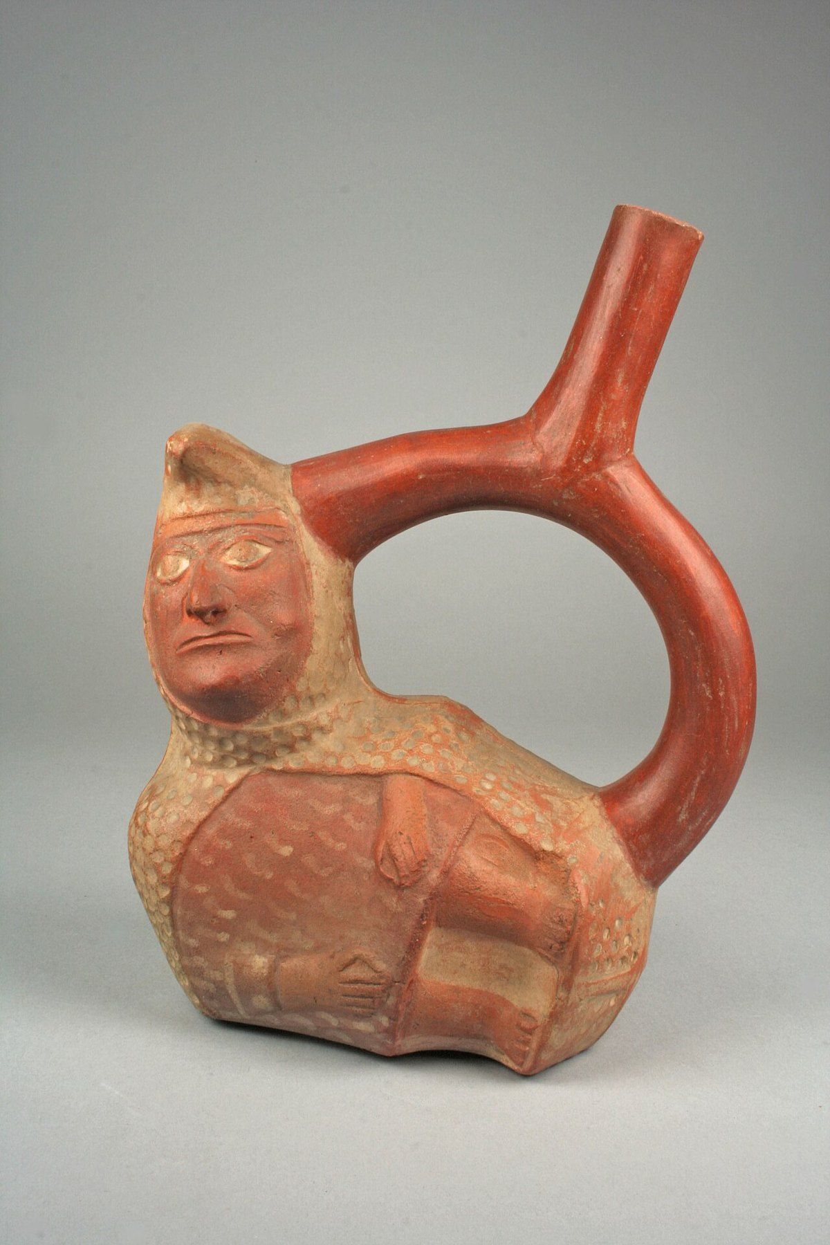 Una vasija de cerámica moche con asa en forma de estribo, decorada con una figura que tiene cuerpo de maní y cabeza y brazos humanos.