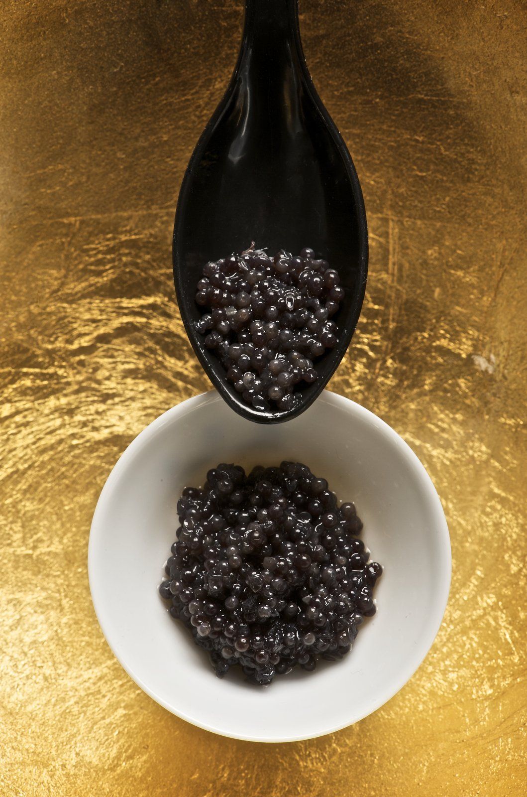 Caviar visto desde arriba: bolitas negras brillantes en una cuchara negra y en un cuenco blanco, sobre un fondo dorado.