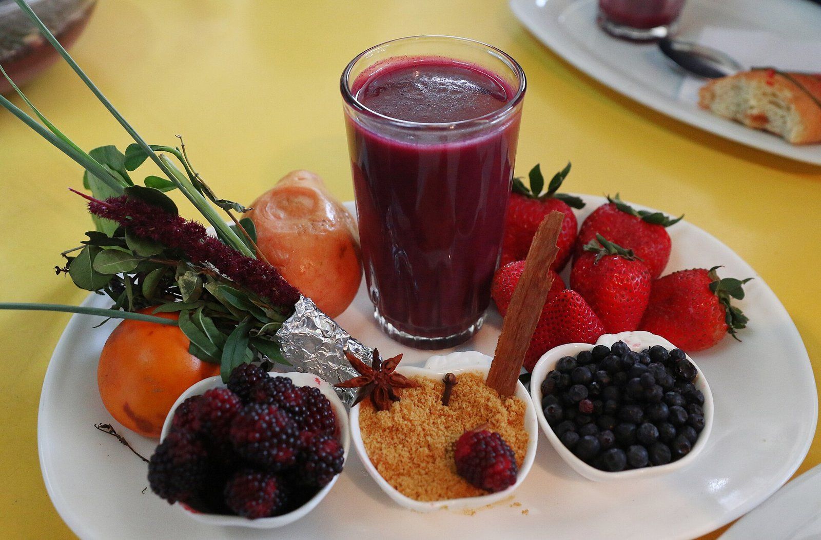 Un vaso de colada morada, una bebida de color violeta, sobre un plato rodeado de frutas, hierbas y especias, entre ellas un palito de canela y una estrella de anís.
