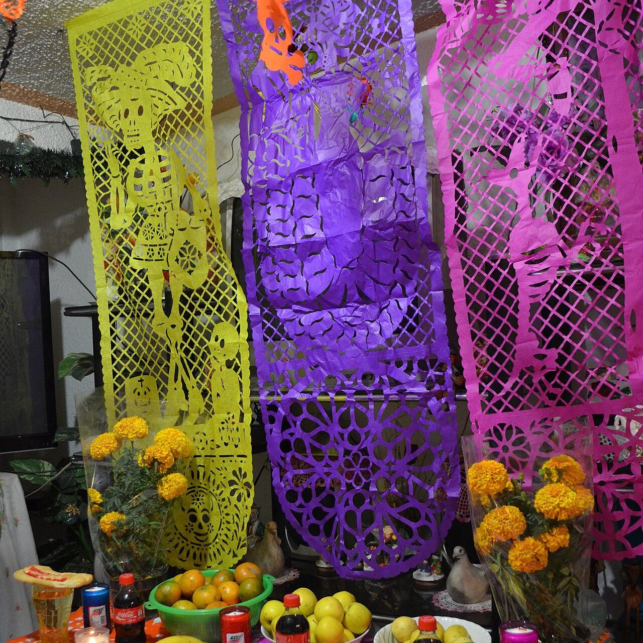 Un altar de Día de Muertos en una casa: papel picado de colores colgado, flores de cempasúchil naranjas, velas, fruta y bebidas sobre un mantel naranja y fucsia.