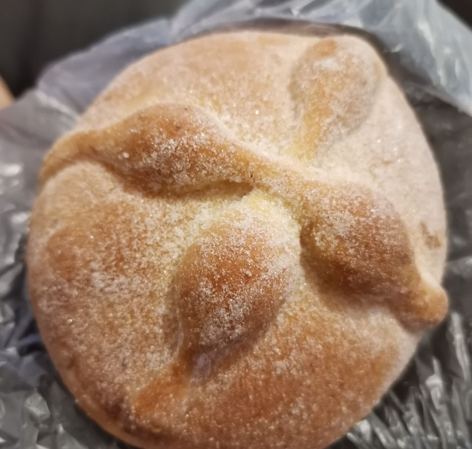 Un pan de muerto visto desde arriba: redondo, dorado, cubierto de azúcar, con tiras de masa con nudos que forman una cruz.