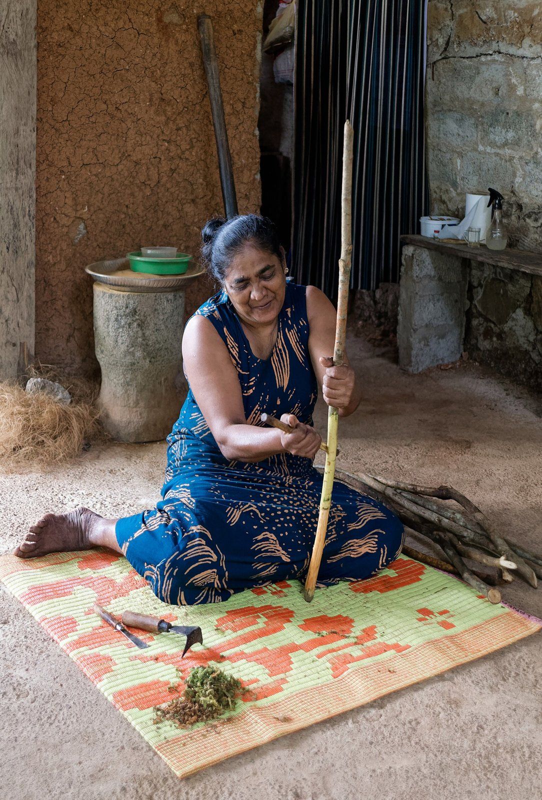 Una mujer sentada en una estera de colores, en el piso de un galpón, trabaja una rama larga y delgada con una herramienta de metal. A su lado hay un atado de ramas cortadas.
